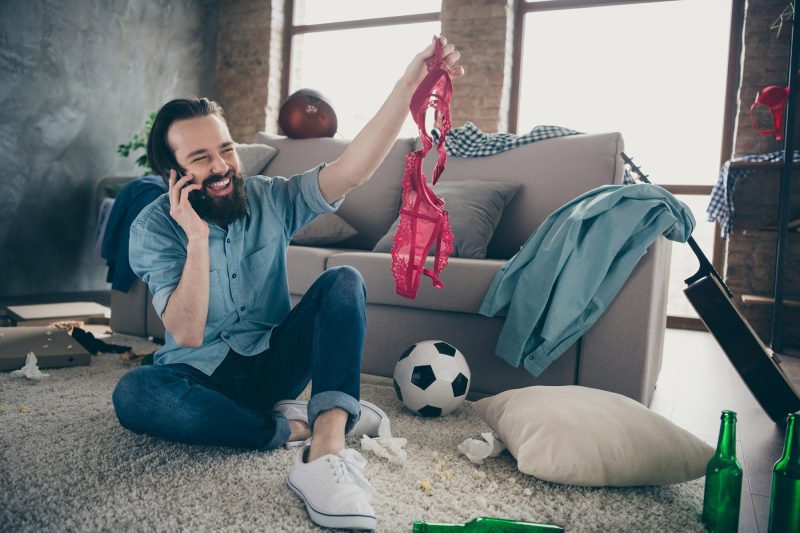 Man in living room on phone holding up a bra and grinning
